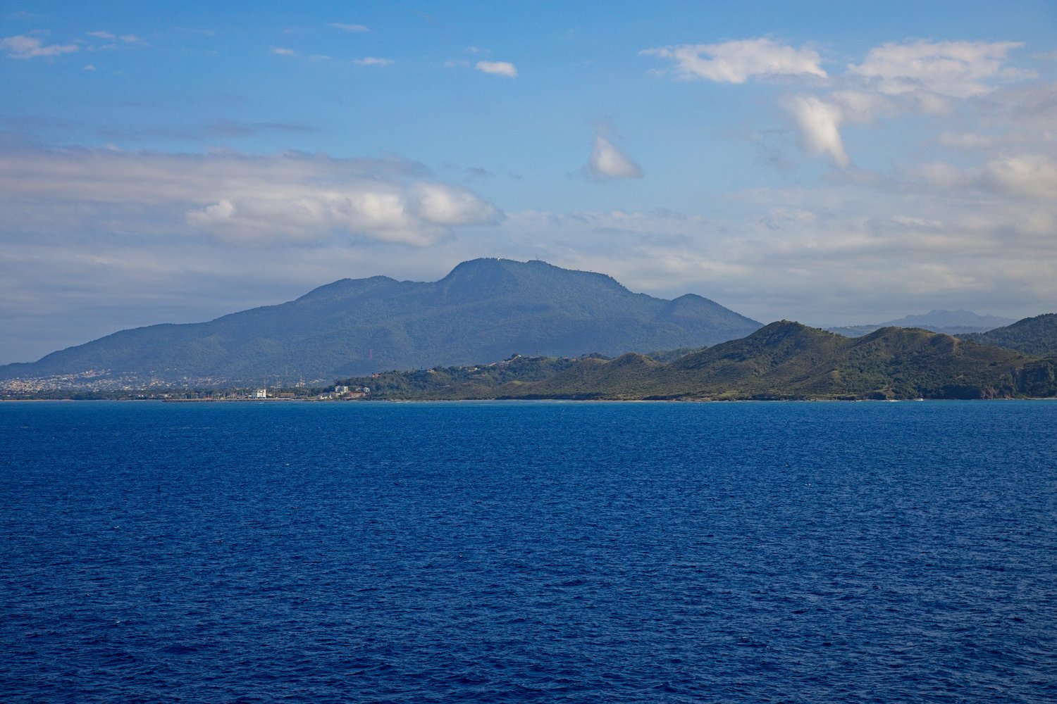A serene lakeshore scene featuring a rugged, exposed shoreline of rocky and muddy terrain in the foreground, dotted with bare, leafless trees along a wooded bank on the left. The calm water stretches out to the horizon where a faint vessel is visible in the distance. Above, a dynamic sky filled with textured clouds in shades of blue and gray suggests a cool, overcast day, likely in late autumn or early spring given the dormant vegetation.